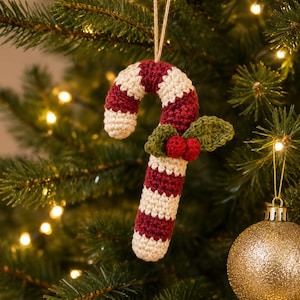 Könnte beinhalten: Ein gehäkelter Zuckerstangen-Anhänger mit abwechselnden horizontalen Streifen aus dunkelrotem und cremefarbenem Garn. Das Ornament ist mit zwei kleinen grünen gehäkelten Blättern und zwei kleinen roten gehäkelten Beeren in der Mitte verziert.