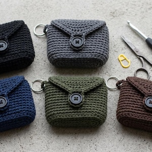 Könnte beinhalten: Fünf kleine, rechteckige, gehäkelte Taschen in Schwarz, Dunkelgrau, Marineblau, Olivgrün und Braun. Jede Tasche hat eine dreieckige Klappe, die mit einem schwarzen Knopf und einem Metallring befestigt ist. Eine Häkelnadel, eine Schere und ein gelber Maschenmarkierer liegen bei.