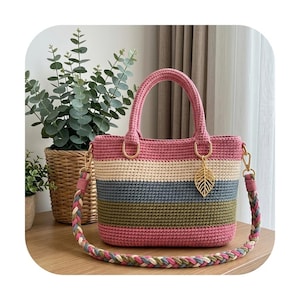 Könnte beinhalten: Eine gehäkelte Handtasche mit horizontalen Streifen in Rosa, Creme, Blau und Olivgrün. Die Tasche hat zwei Tragegriffe und einen abnehmbaren, geflochtenen Schulterriemen in passenden Farben. Ein goldfarbener Metallanhänger in Blattform hängt an einem Ring.
