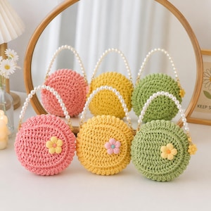 Könnte beinhalten: Drei runde Häkeltaschen in Pink, Gelb und Grün. Jede Tasche verfügt über ein strukturiertes Strickmuster, einen einzelnen Henkel aus weißen Kunstperlen und eine kleine, einfarbige Blumenverzierung, die mittig auf der Vorderseite angebracht ist.