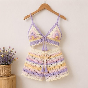 Könnte beinhalten: Ein zweiteiliges Häkelset bestehend aus einem Triangel-Bralette-Top und passender Shorts. Das Kleidungsstück zeigt ein Streifenmuster in Pastelltönen wie Lavendel, Hellgelb und Creme, abgerundet mit gewellten Kanten und Lavendelquasten.