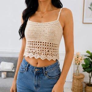 Könnte beinhalten: Ein beiges Häkel-Crop-Top mit quadratischem Ausschnitt, dünnen Trägern und einem gewellten Saum. Der obere Teil besteht aus einem dichten, festen Maschenmuster, während der untere Teil ein offenes, luftiges Gitterdesign aufweist. Kombiniert mit einer blauen Jeans.