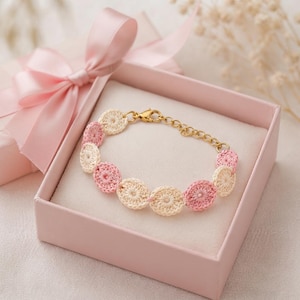 Könnte beinhalten: Ein Armband mit abwechselnden cremefarbenen und hellrosa gehäkelten kreisförmigen Motiven, die durch eine goldfarbene Metallkette verbunden sind. Das Armband hat einen goldfarbenen Karabinerverschluss und eine Verlängerungskette in einer rosa Schachtel.