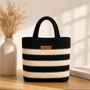 Könnte beinhalten: Eine schwarz-cremefarbene, gestreifte Häkeltasche mit zwei Tragegriffen. Die Tasche hat ein rechteckiges, braunes, lederartiges Etikett in der Mitte der Vorderseite, auf dem das Wort Handmade eingeprägt ist. Das Häkelmuster ist durchgehend.