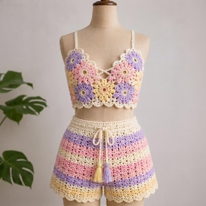 Könnte beinhalten: Ein zweiteiliges Häkelset bestehend aus einem bauchfreien Oberteil und einer Shorts. Das Oberteil zeigt ein florales Granny-Square-Muster in Pastellgelb, Rosa und Lavendel. Die Shorts haben horizontale Streifen in denselben Farben mit Kordelzug und Quasten.