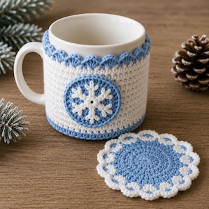 Könnte beinhalten: Ein weißer Keramikbecher mit einem abnehmbaren Häkelüberzug in Weiß und Hellblau, verziert mit einem zentralen weißen Schneeflockenmotiv auf blauem kreisförmigem Hintergrund, dazu ein passender runder Häkeluntersetzer mit gewelltem Rand.