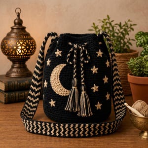 Könnte beinhalten: Eine schwarze Häkel-Beuteltasche mit einem beigefarbenen Halbmond- und Sternenmuster. Die Tasche verfügt über einen Kordelzug mit zwei geflochtenen Schnüren, die in Quasten enden, sowie einen breiten Schultergurt mit beigem und schwarzem Zickzackmuster.