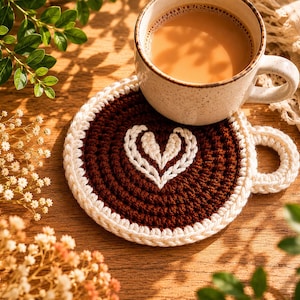 Könnte beinhalten: Eine beigefarbene Kaffeetasse steht auf einem handgefertigten Häkeluntersetzer. Der Untersetzer ist braun mit einem weißen Herzmuster und einem weißen Rand. Die Tasse enthält Kaffee. Der Untersetzer hat einen Durchmesser von ca. 10 cm.