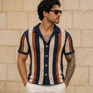 Puede incluir: Una camisa de manga corta con botones y un patrón de rayas verticales en azul marino, crema y naranja óxido. La prenda está confeccionada con un tejido de ganchillo texturizado de punto abierto, con un cuello clásico y cuatro botones marrones.