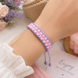 Könnte beinhalten: Ein gewebtes Freundschaftsarmband mit einem sich wiederholenden Muster aus violetten, rosafarbenen und weißen Fäden. Das Armband hat ein flaches, geflochtenes Design mit zwei verstellbaren violetten Kordeln, die jeweils mit einem Knoten enden.