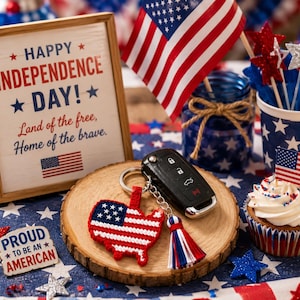 Puede incluir: Un llavero con un mapa de los Estados Unidos tejido en rojo, blanco y azul, unido a una llave de coche negra y una borla multicolor. El llavero descansa sobre una rodaja de madera junto a un cartel que dice Happy Independence Day.