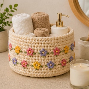 Könnte beinhalten: Ein runder, cremefarbener Häkelkorb mit zwei horizontalen Reihen kleiner, bunter Blumenapplikationen in Rosa, Gelb und Violett mit grünen Blättern. Der Korb enthält mehrere gerollte weiße und hellbraune Handtücher sowie einen weißen Seifenspender.