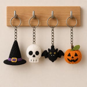 Könnte beinhalten: Vier gehäkelte Schlüsselanhänger hängen an einer Holzleiste. Die Anhänger bestehen aus einem schwarzen Hexenhut mit violettem Band, einem weißen Totenkopf, einer schwarzen Fledermaus mit gelben Augen und einem orangefarbenen Kürbis mit grünem Blatt.