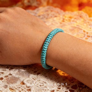May include: A turquoise bracelet with a woven design is worn on a wrist. The bracelet is a bright, solid color and appears to be made of a flexible material. The background is a soft, warm color.