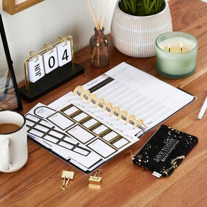 K&ouml;nnte beinhalten: Ein Schreibtisch-Setup mit einem Planer, Aufklebern und einem kleinen schwarzen Beutel mit goldenen Akzenten. Ein goldfarbener Schreibtischkalender zeigt den 04. Juni. Eine brennende Kerze und eine Tasse Kaffee sind ebenfalls vorhanden.