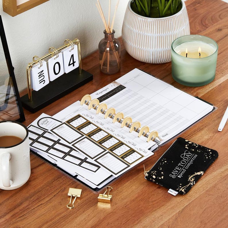 K&ouml;nnte beinhalten: Ein Schreibtisch-Setup mit einem Planer, Aufklebern und einem kleinen schwarzen Beutel mit goldenen Akzenten. Ein goldfarbener Schreibtischkalender zeigt den 04. Juni. Eine brennende Kerze und eine Tasse Kaffee sind ebenfalls vorhanden.
