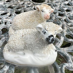 May include: A ceramic figurine featuring two sheep with textured, granular off-white coats. The top sheep has a pinkish nose, while the bottom sheep has a black nose. Both figures have painted eyes and ears, resting on a smooth, white base.