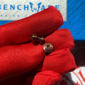 May include: Two small, metallic, cylindrical hardware components with a ribbed, circular top surface. These fasteners feature a central shaft and a stepped, concentric design, held against a bright red fabric background with a Benchmade logo visible.
