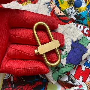 May include: A gold-colored metal clasp with a rectangular bar in the center. The clasp is in the shape of a U. The background is a red fabric and a comic book print.