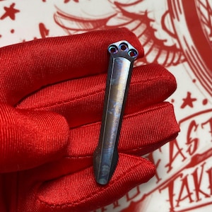 May include: A small, metallic tool with a unique design. The tool has a rectangular body with a series of three circular holes at the top. The metal has a gradient of colors, including blue and purple. The tool is held in a red glove.