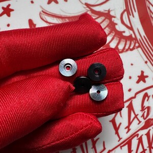 Puede incluir: Cuatro objetos peque&ntilde;os y circulares. Dos son plateados con una superficie estampada y dos son negros. Los objetos descansan sobre una superficie roja texturizada. El fondo presenta dise&ntilde;os gr&aacute;ficos rojos y blancos.