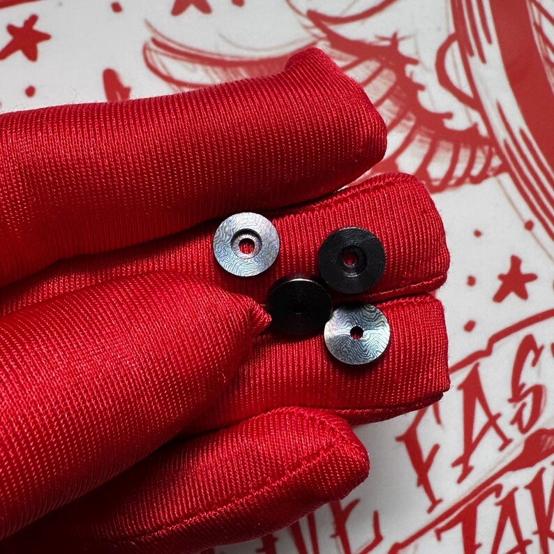 Puede incluir: Cuatro objetos peque&ntilde;os y circulares. Dos son plateados con una superficie estampada y dos son negros. Los objetos descansan sobre una superficie roja texturizada. El fondo presenta dise&ntilde;os gr&aacute;ficos rojos y blancos.