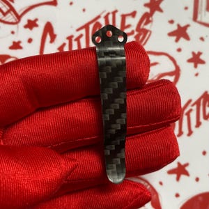 May include: A carbon fiber pocket clip with a textured, woven pattern. The clip has three holes at the top and a rounded bottom. The clip is held against a red fabric background with a white and red design.