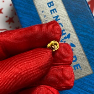 May include: A small, gold-colored pin with a spiral design. The pin is held against a red fabric background, with a blue and white box in the background. The pin has a circular design with concentric circles.