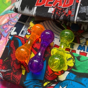 May include: Three colorful, glitter-filled glass wands in orange, purple, and green. The wands have a dumbbell shape with a rounded top and bottom, and a narrow center. The wands are set against a comic book fabric background.