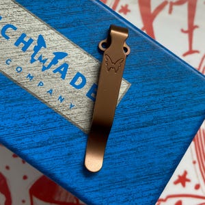 May include: A copper-colored pocket clip with a butterfly logo, resting on a blue box with the text "Benchmade Company". The clip is made of metal and has a smooth, reflective surface. The butterfly logo is etched into the clip.