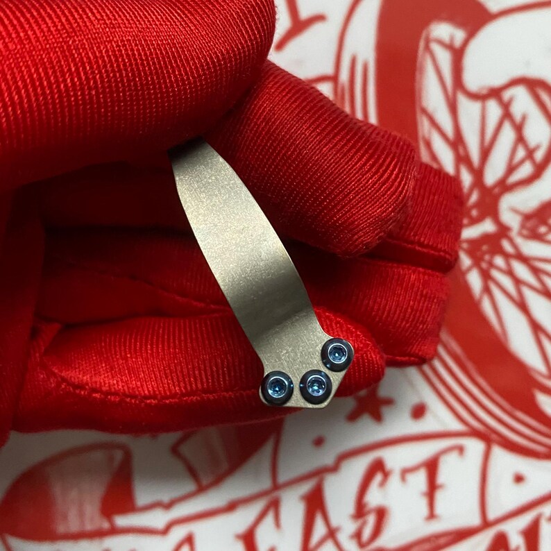 May include: A metallic silver pocket clip with three small blue screws. The clip is held against a red fabric background with a white and red graphic design in the background.