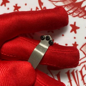 Puede incluir: Primer plano de un peque&ntilde;o clip met&aacute;lico plateado con tres tornillos negros. El clip se sostiene contra una tela roja vibrante. El fondo presenta una superficie blanca con dise&ntilde;os de estrellas y texto rojos.