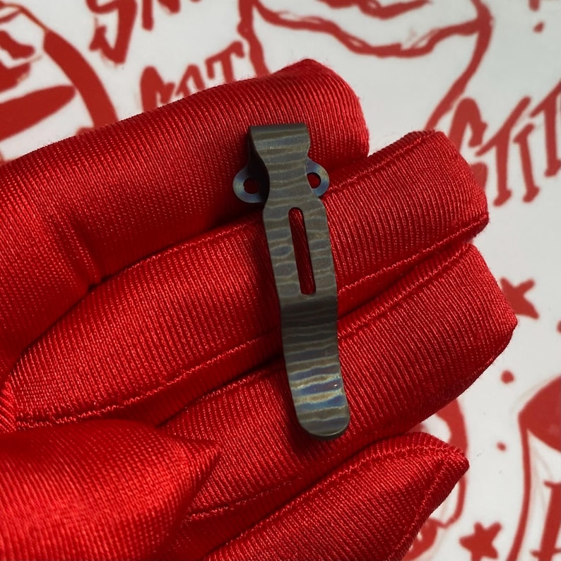 May include: A small, patterned metal clip with a rectangular cutout and two circular holes at the top. The clip has a textured, layered appearance with blue and gray tones. The clip is held against a red fabric background.