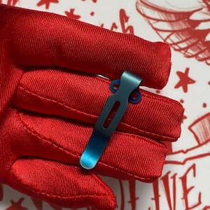 May include: A teal-colored pocket clip with a rectangular cutout and two circular mounting holes. The clip is held against a red fabric glove. The background features a white surface with red decorative elements.