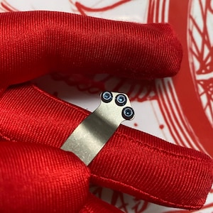 May include: A close-up of a small, silver-colored metal object with three small, blue-colored circular accents. The object is set against a vibrant red background, creating a striking contrast. The image is well-lit, highlighting the details of the metal and blue accents.