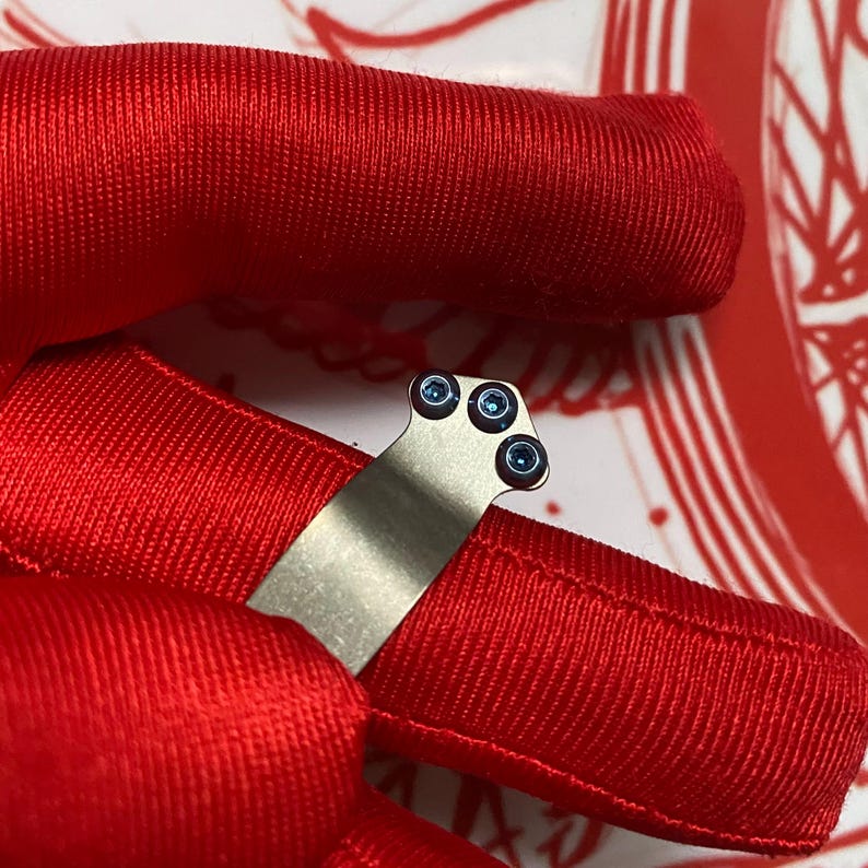 May include: A close-up of a small, silver-colored metal object with three small, blue-colored circular accents. The object is set against a vibrant red background, creating a striking contrast. The image is well-lit, highlighting the details of the metal and blue accents.