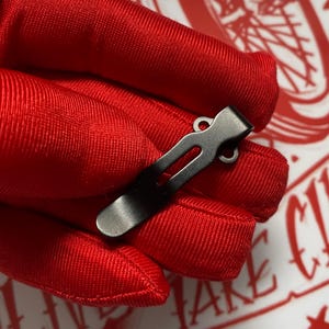 May include: A dark gray metal pocket clip with a curved end and a slotted design. The clip is shown against a red background.