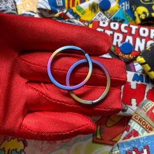 May include: Two iridescent, hinged hoop earrings. The earrings display a spectrum of colors, including blue, purple, and gold. The earrings are held in a red glove, with comic book patches in the background.