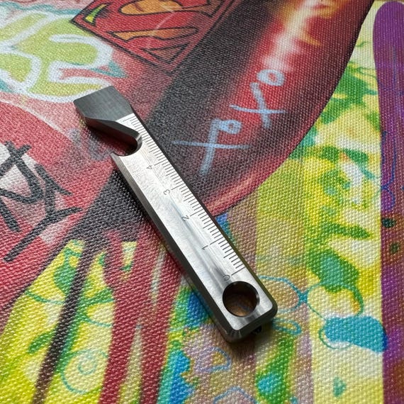 A high-polished titanium multitool ruler that's also a pry bar, bottle opener, and glass breaker — custom-made EDC that looks like it belongs in a sci-fi movie.
