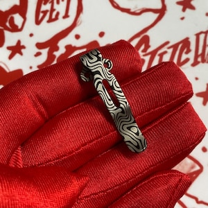 May include: A silver and black patterned pocket clip. The clip has a curved hook and a long, slender body with a wavy, organic design. The clip is held against a red fabric background.