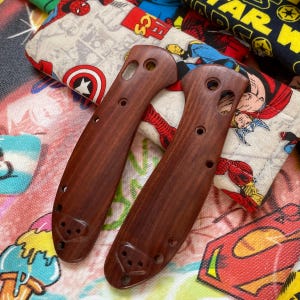 May include: Two wooden knife handles with a rich, reddish-brown hue. Each handle features a series of drilled holes and a decorative carved design at the base. The handles are displayed against a colorful fabric background with comic book characters.