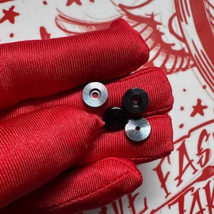 May include: Close-up of four small, circular objects. Two are silver with a patterned design, and two are black. The objects are resting on a red, textured surface. The background includes a red and white design.