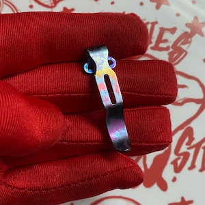 May include: A metal pocket clip featuring an iridescent, multi-colored finish with shades of blue, purple, yellow, and pink. The clip has a curved, elongated shape with a central vertical slot and two circular mounting holes at the top end.