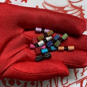May include: A collection of colorful metal valve stem caps in various colors, including red, gold, blue, and green. Each cap has a textured finish and is connected to a black rubber base. The caps are displayed on a red fabric surface.