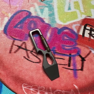 May include: A gray multi-tool with a spring-loaded mechanism and a rectangular blade. The tool has a carabiner-style top and a small hole. The background is a colorful graffiti design with pink, blue, green, and white paint.