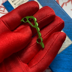 May include: A small, bright green metal tool with a unique, open design. The tool has a series of holes and curves, held against a red fabric background. The tool appears to be a specialized accessory.