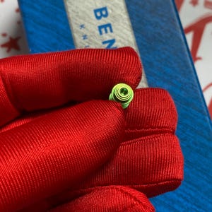 May include: A small, green, spiral-shaped object with a central green gem is held in a red fabric. The object is set against a blue and white background with the word "BEN" visible.