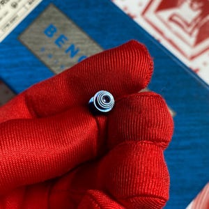 May include: A small, blue metal object with concentric circles and a central hole. The object is held in a red fabric. The word "BEN" is partially visible in the background on a blue and gray surface.