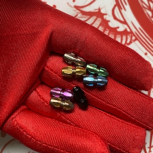 May include: A collection of small, cylindrical metal beads featuring faceted surfaces in various metallic finishes including silver, gold, green, blue, purple, and black. Each bead has a central groove and a polished, reflective texture.