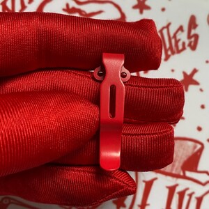 May include: A vibrant red metal clip with a rectangular cutout and two small holes at the top. The clip is held against a background of red fabric. The clip's design suggests it is a functional accessory, possibly for securing items.